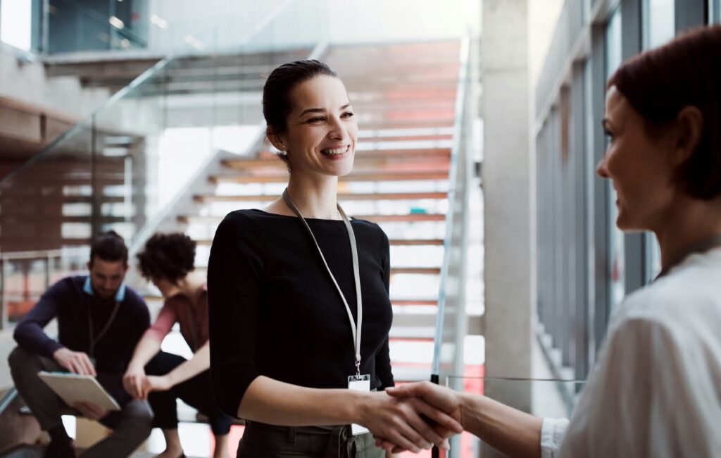 A photo of businesswomen shaking hands in the foreground whilst in the background, out-of-focus, a man and a woman review something on what looks like a clipboard, or possibly a large tablet.