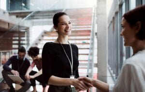 A photo of businesswomen shaking hands in the foreground whilst in the background, out-of-focus, a man and a woman review something on what looks like a clipboard, or possibly a large tablet.